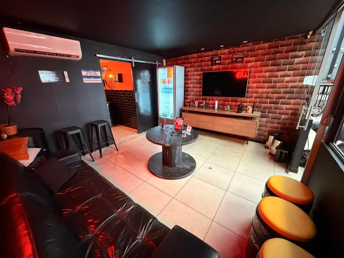 Sala de espera de oficina automotiva com sofá preto, mesa redonda, TV, geladeira de bebidas e bancos estilo tambor