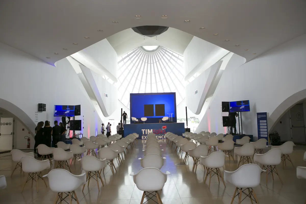 Sala de evento com cadeiras brancas alinhadas voltadas para palco azul com tela, em ambiente arquitetônico branco e iluminado