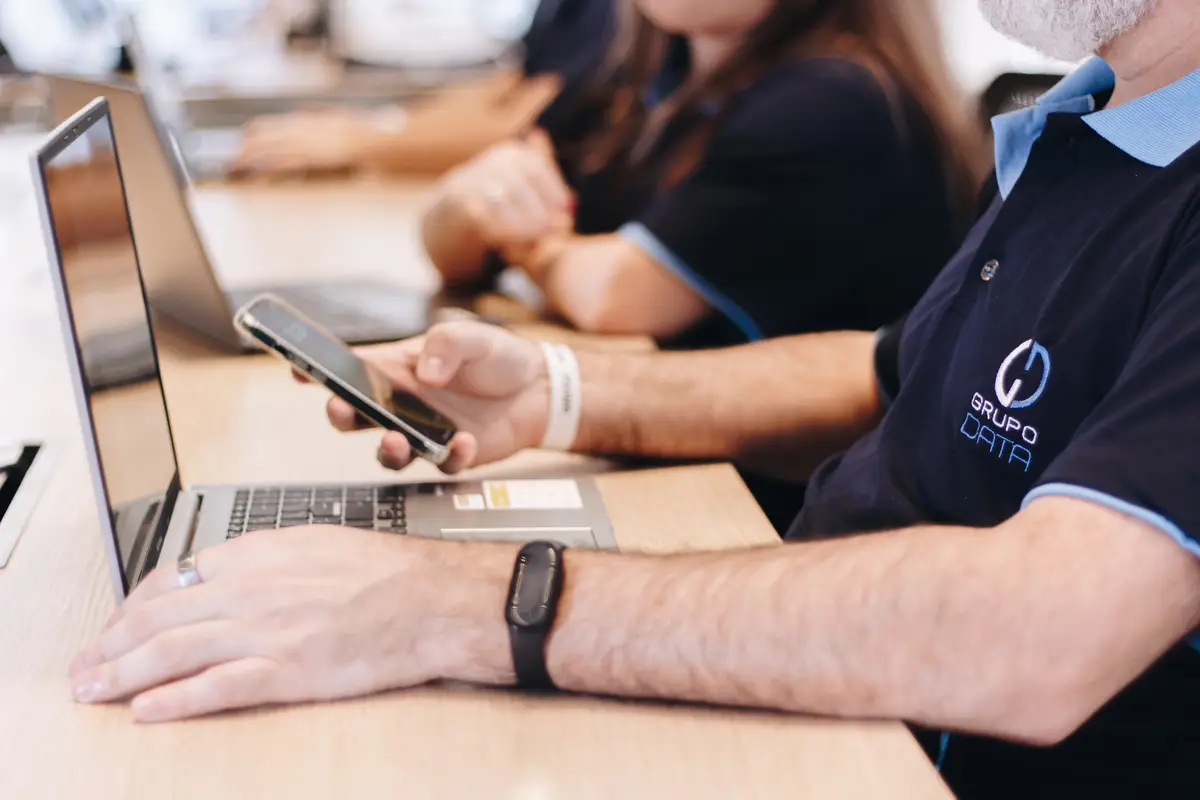 Pessoa usando smartphone e laptop na mesa com camiseta do Grupo Data
