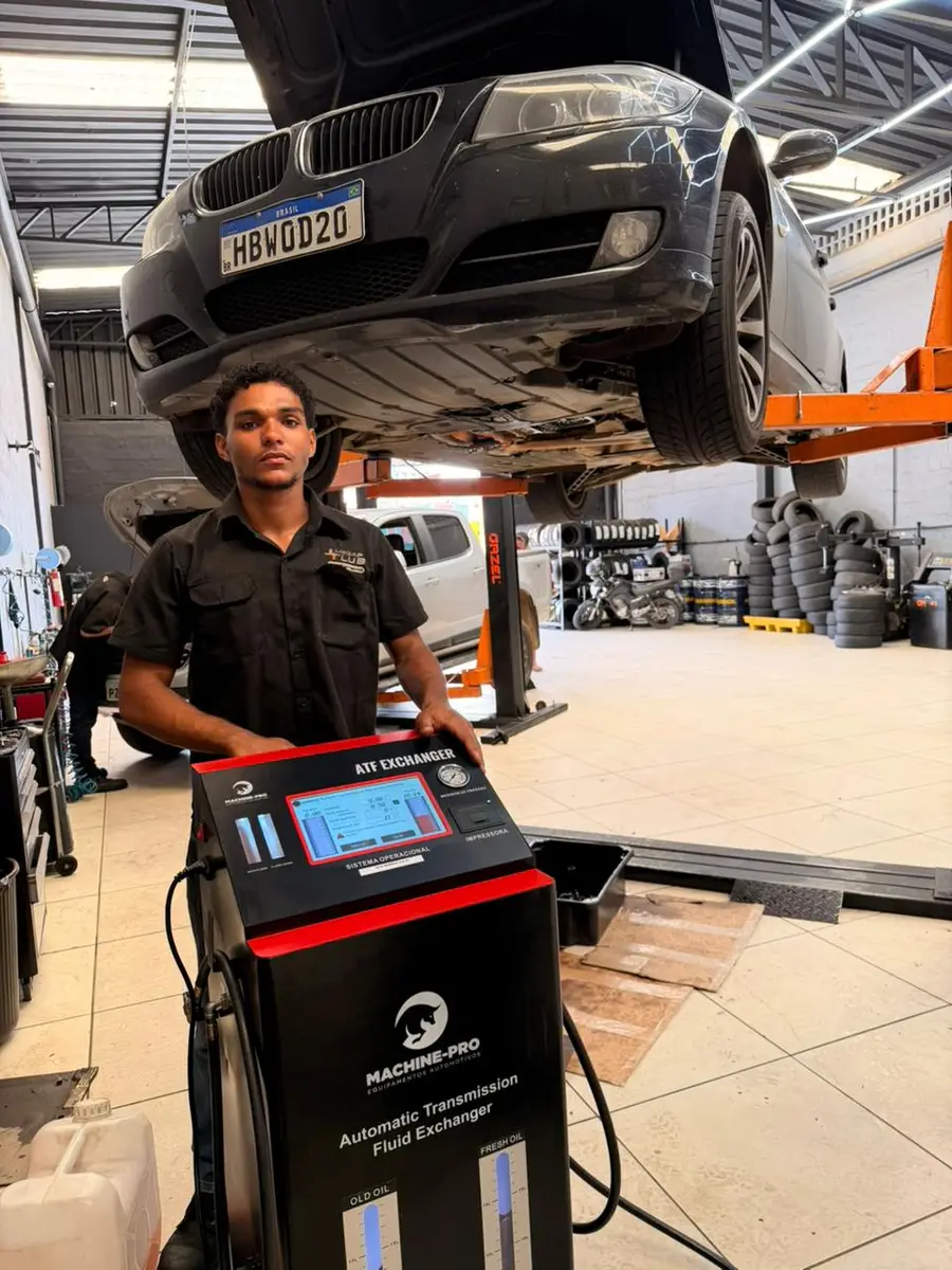 Técnico automotivo operando máquina de troca de óleo de câmbio automático em oficina com carro elevado no elevador