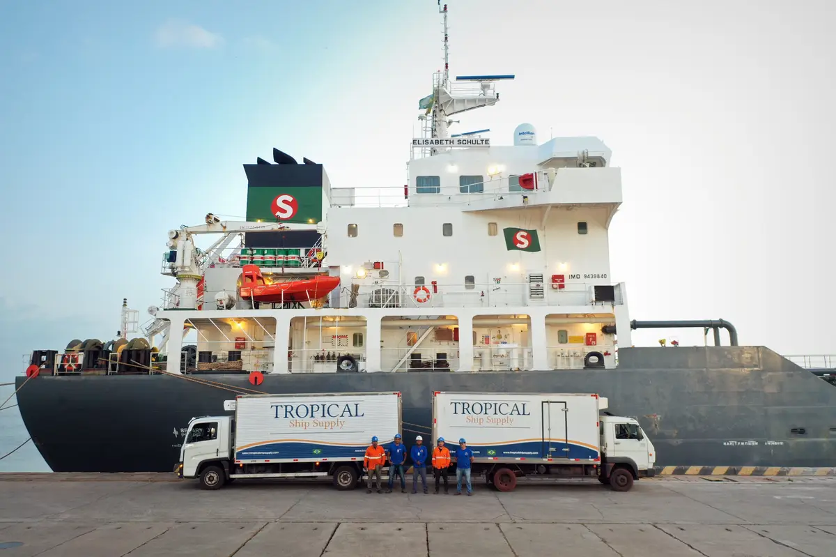 Two Tropical Ship Supply trucks parked beside a large cargo ship with workers standing between them