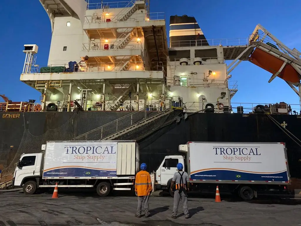 Two Tropical Ship Supply trucks parked by a docked cargo ship with workers standing nearby