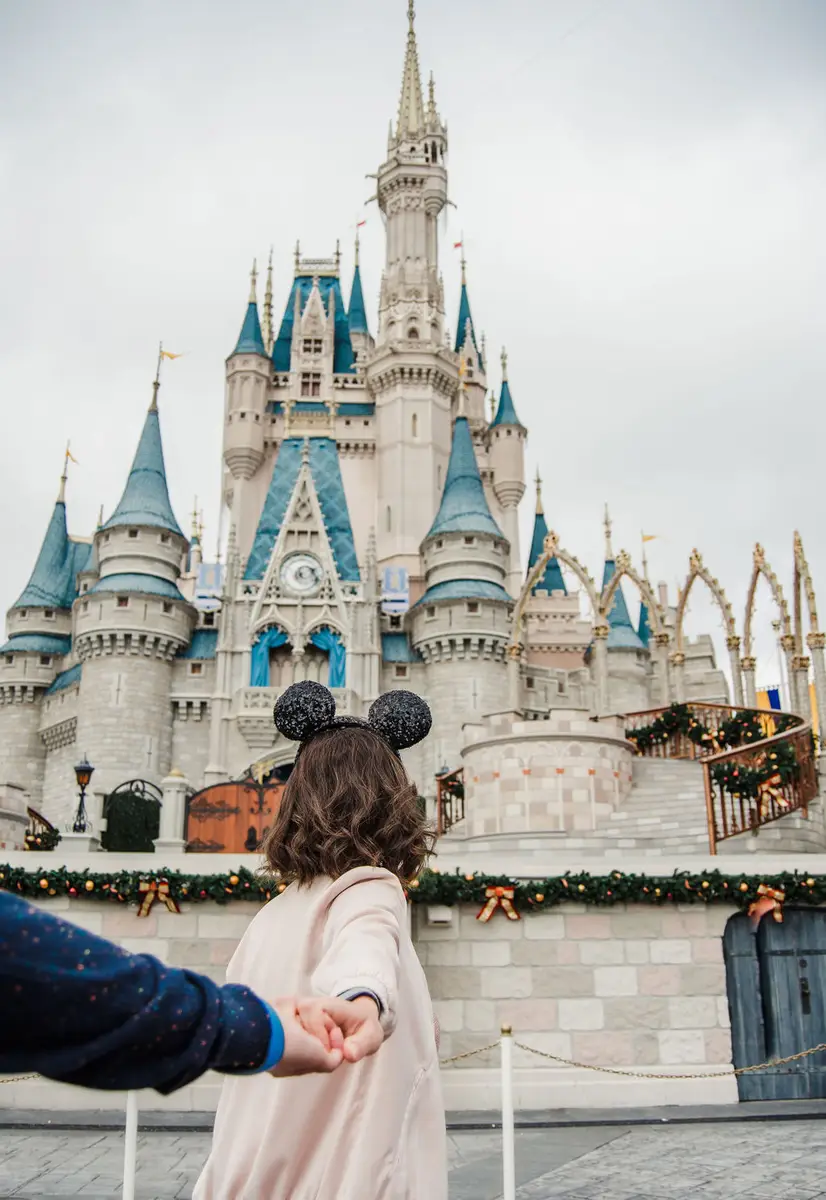 Pessoa de costas com tiara de orelhas de Mickey segurando a mão de outra pessoa em frente ao castelo da Cinderela na Disney