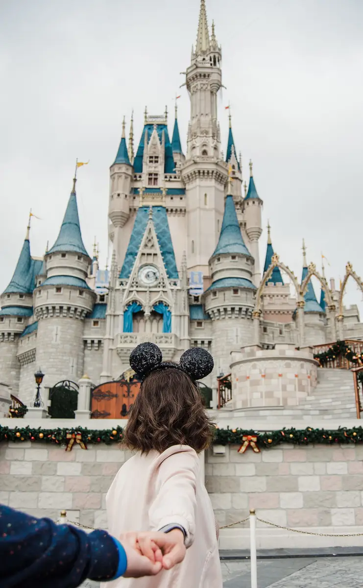 Pessoa de costas usando tiara de orelhas da Minnie segurando a mão de outra pessoa em frente ao castelo da Cinderela na Disney
